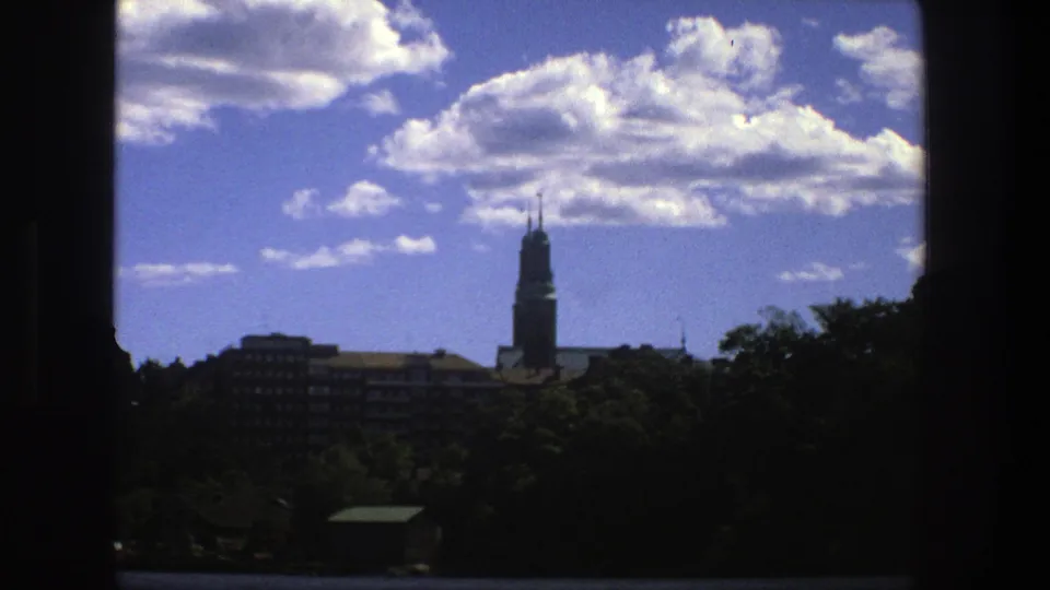 Final 1975 frame of a Stockholm skyline, with the spire offset right, wooded waterfront foreground, and bright cumulus clouds.