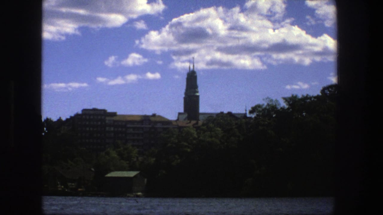 1975 Stockholm boat-view frame showing water, shaded shoreline trees, apartment buildings, and a tall church spire beneath a blue sky.