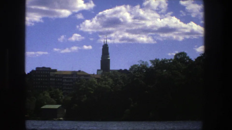 1975 Stockholm waterfront view from a boat, with dark vertical framing around trees, apartment blocks, and a distant spire.