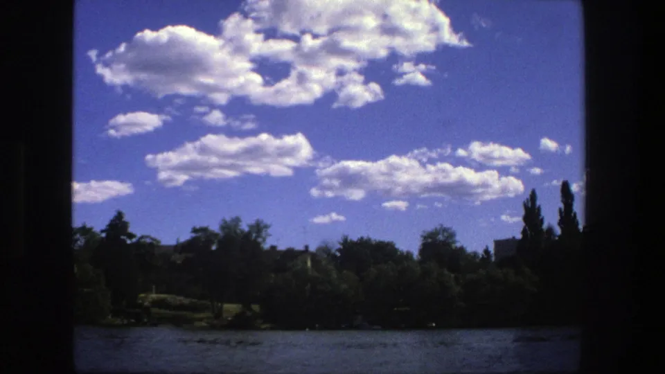 1975 Stockholm boat-view frame with bright clouds above a green shoreline, water, and low urban structures in the distance.