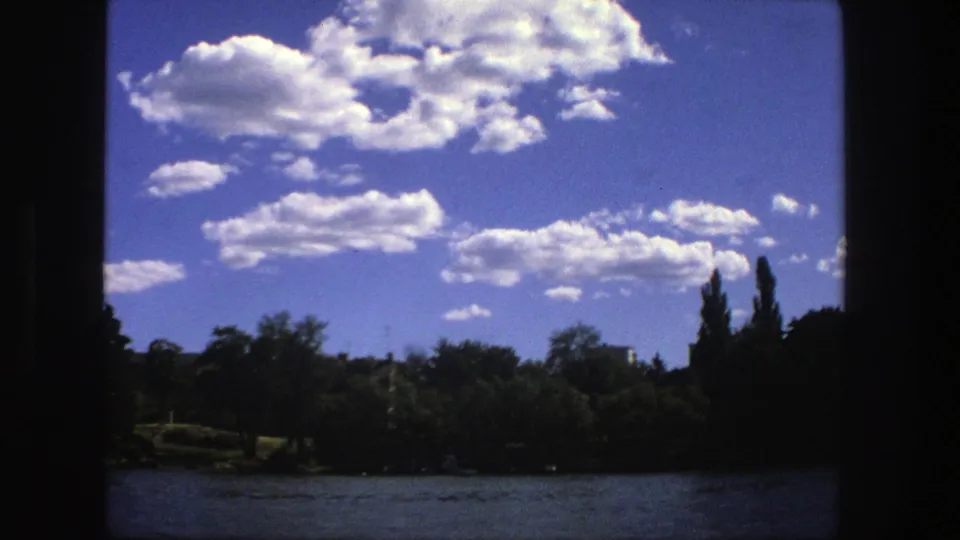 1975 Stockholm waterfront from a boat, showing bright summer clouds, trees along the shore, water, and distant low buildings.