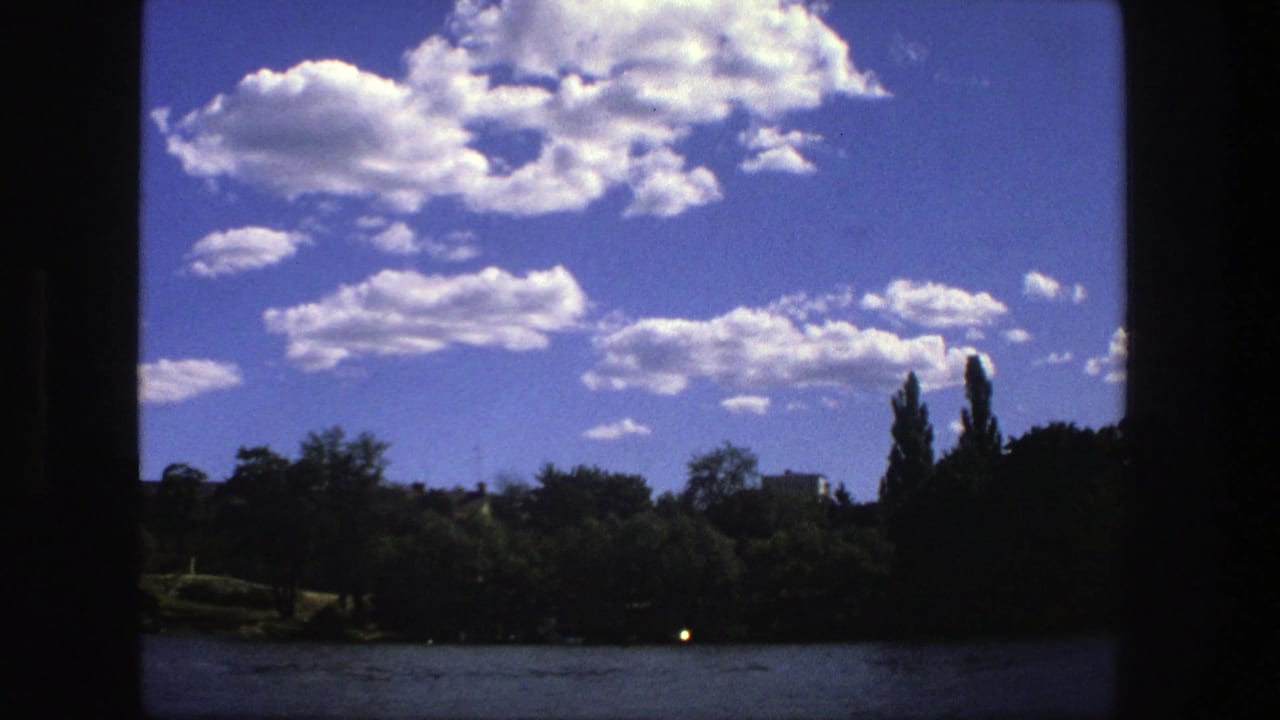 1975 Stockholm home-movie view across a lake toward trees under large cumulus clouds, framed by dark 8mm borders.