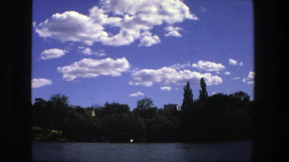 A steady 8mm view across Stockholm lake water to a shaded treeline beneath bright clouds and blue sky.