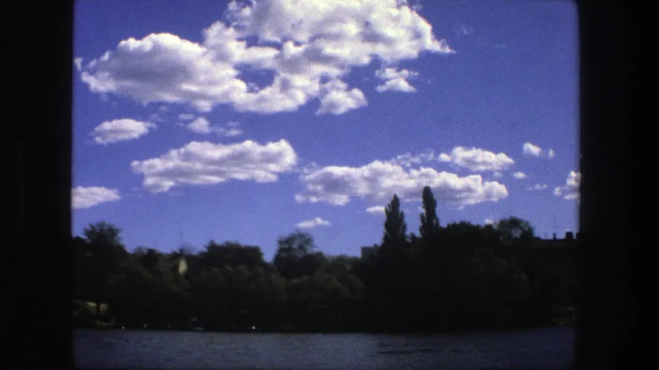 1975 8mm Stockholm lakeside view with low water, silhouetted trees, tall poplars, and bright cumulus clouds.