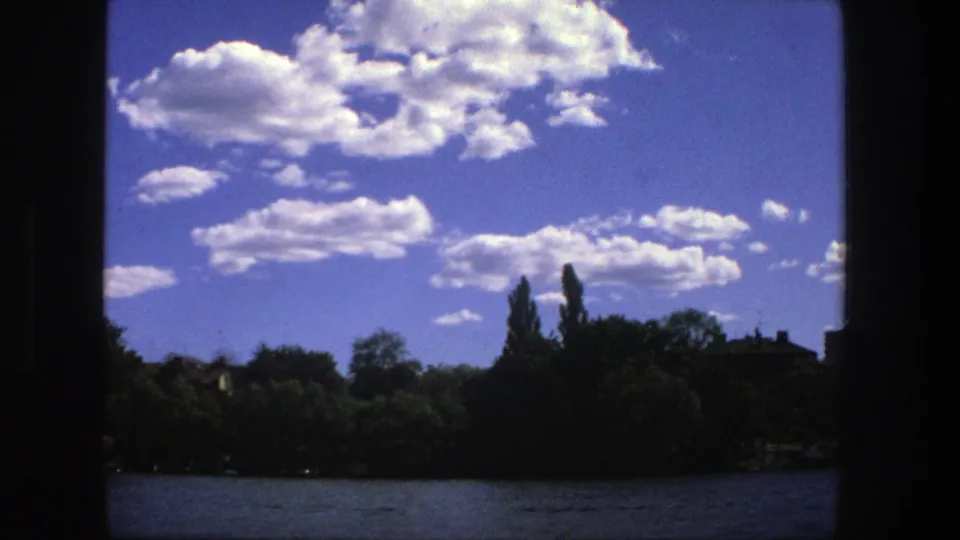 Stockholm 1975 home-movie frame showing a calm lake foreground, dark treed shore, and bright cloud formations.