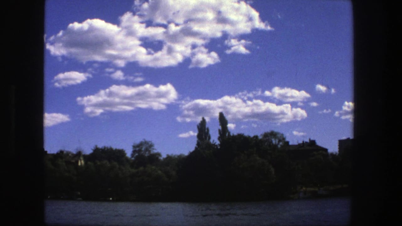 1975 Stockholm waterfront view with bright clouds, dark foreground framing, trees, water, and distant buildings.