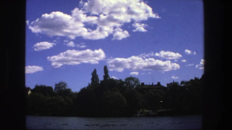 1975 Stockholm shoreline scene with open water, leafy trees, distant rooftops, and scattered white clouds.