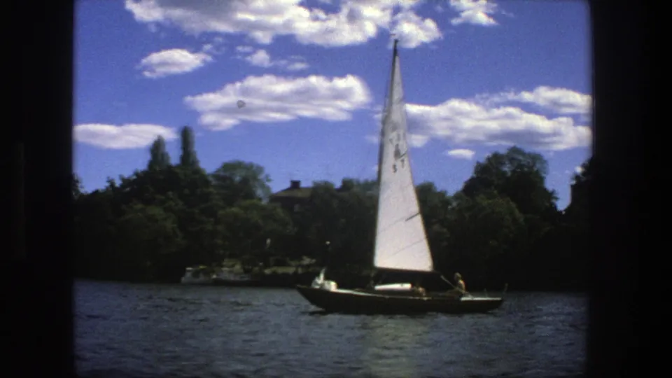 1975 Stockholm waterfront view with a small sailboat, visible occupants, treelined shore, and bright clouds.