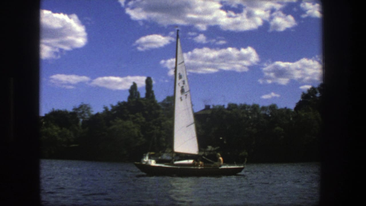 White-sailed boat crossing dark water beneath a blue sky with scattered clouds.