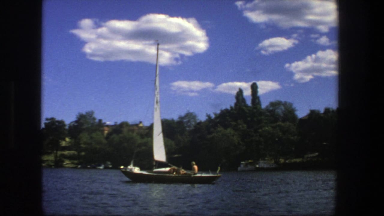 Small sailboat farther across the river with trees, buildings, and clouds behind it.