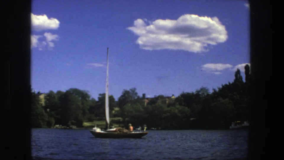A 1975 Stockholm waterfront frame shows the sailboat receding across the water with trees and clouds behind it.