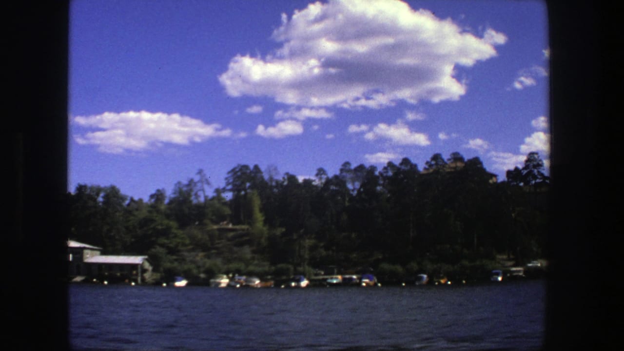 1975 Stockholm home-movie frame showing rippled water, small boats along a wooded shore, and bright clouds in a blue sky.