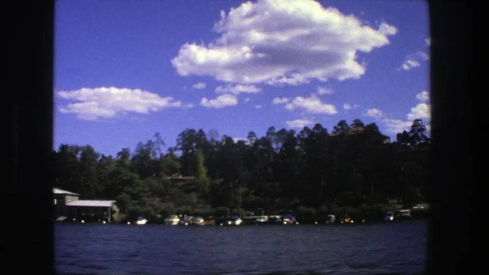 1975 Stockholm home-movie frame showing rippled water, small boats along a wooded shore, and bright clouds in a blue sky.