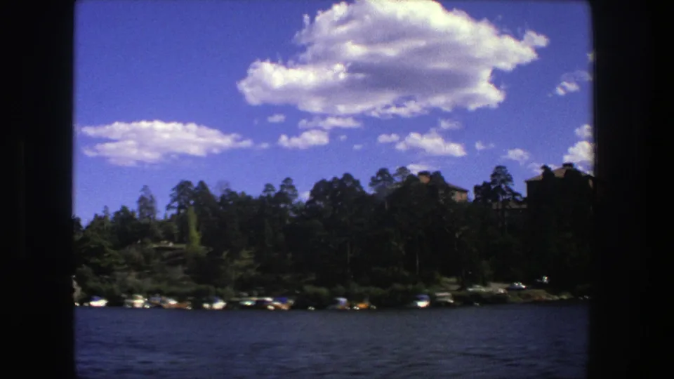 1975 Stockholm home-movie frame showing a moving view of dark water, shoreline boats, dense trees, and large white clouds.