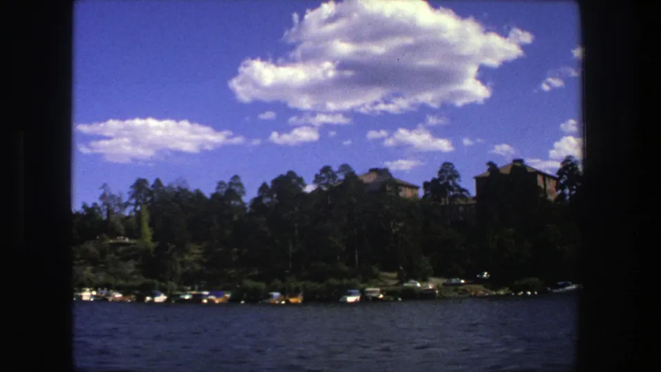 1975 Stockholm frame from a boat, with open water in front of a tree-lined shore, small boats, and a partly visible building.