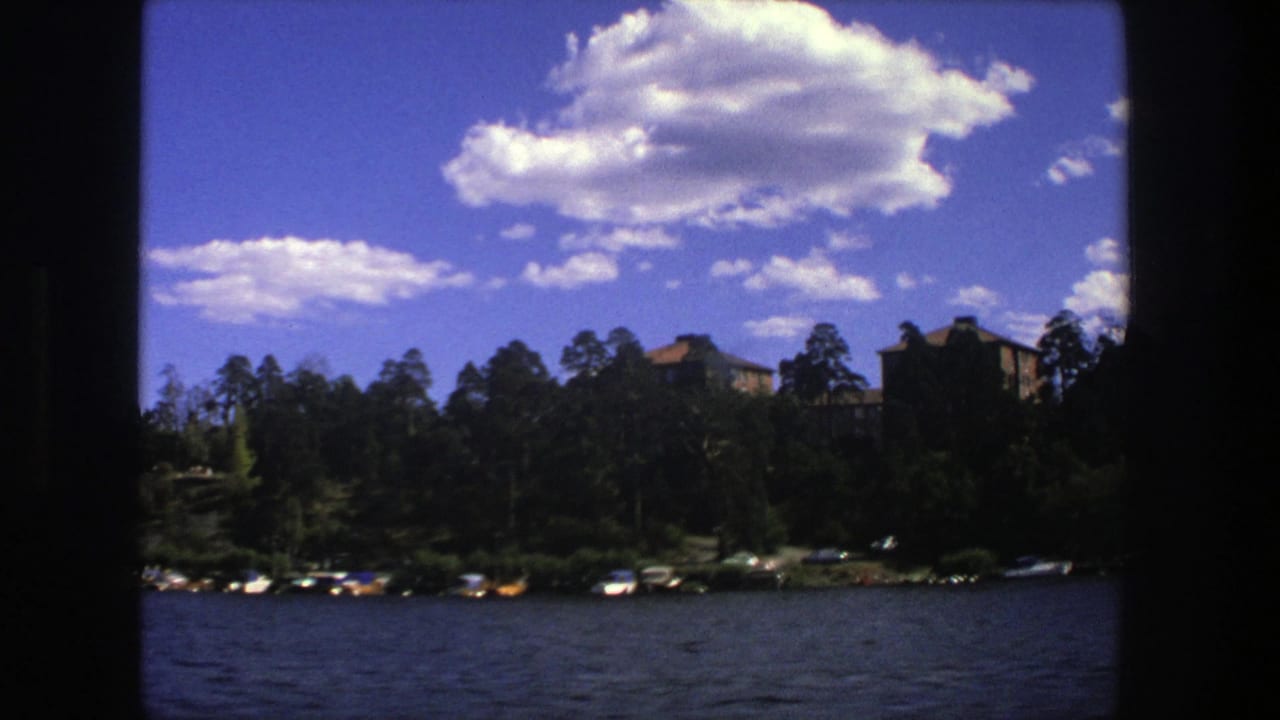1975 Stockholm lake view with small boats, dark foreground borders, trees, distant buildings, and bright clouds over blue water.
