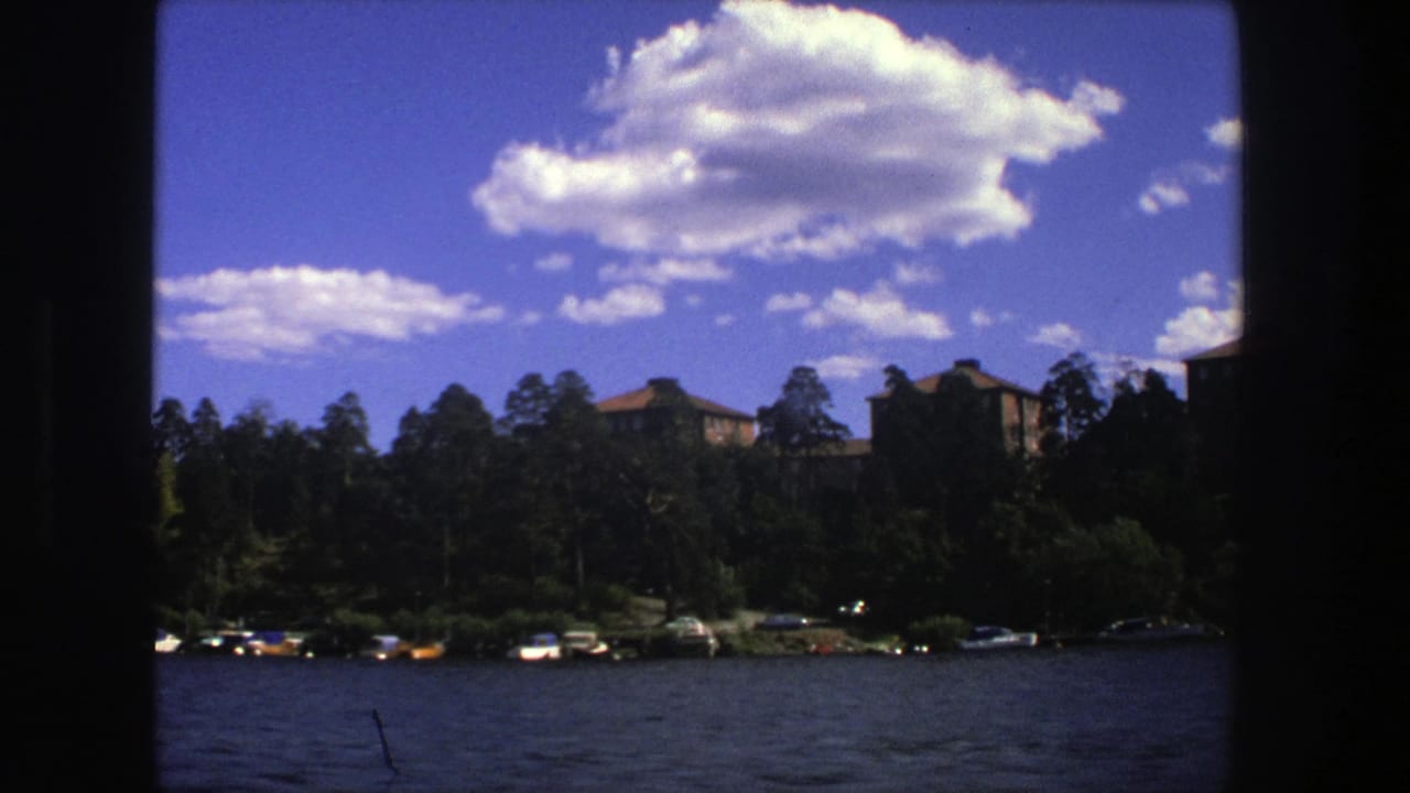 Wide lakeside view with shoreline boats, trees, buildings, and bright cloud formations.