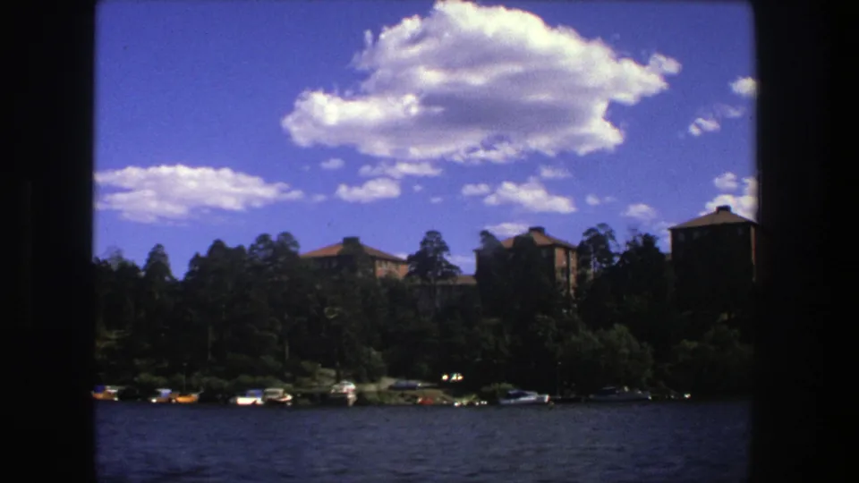 1975 Stockholm lakeside frame showing dark film borders, rippled water, shoreline boats, trees, and blocky buildings in the distance.