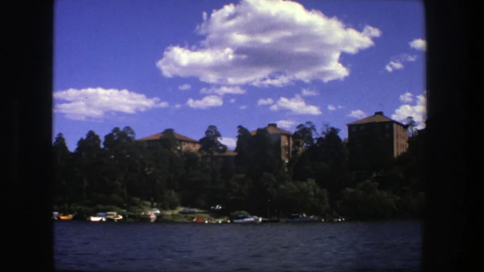 1975 Stockholm lake scene with small boats at the wooded shore, distant buildings, dark frame edges, and a bright cloud-filled sky.