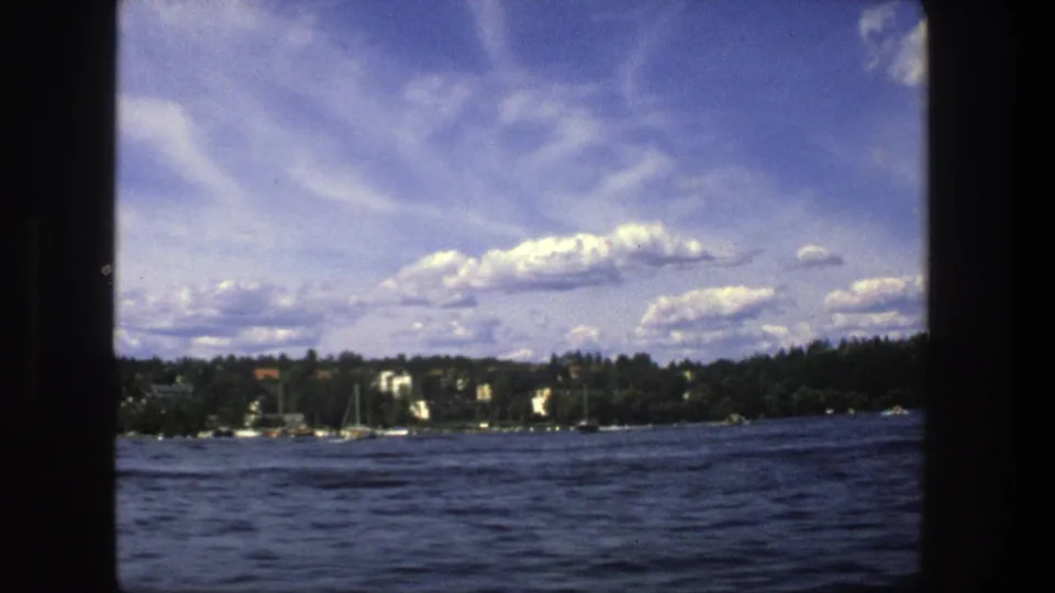 1975 Stockholm lakeside view across dark water toward wooded shore, small boats, sailboat masts, and distant light-colored buildings.