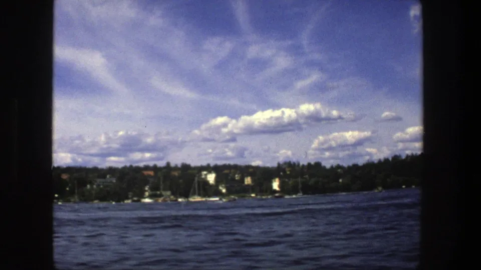 1975 Stockholm boat-perspective frame with rippled lake water, distant shoreline, moored boats, trees, and a cloud-filled sky.
