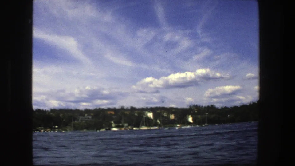1975 Stockholm waterfront frame showing dark lake water, low wooded shore, small boats, pale buildings, and bright scattered clouds.