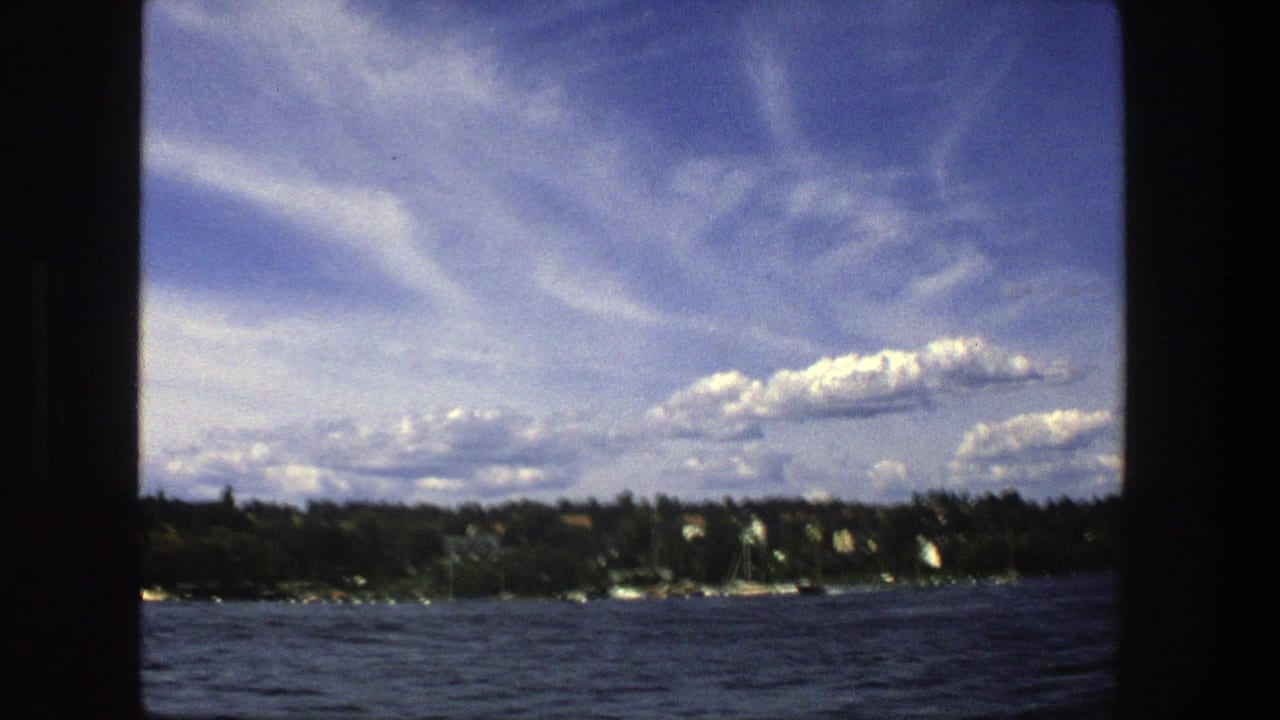 Distant shoreline, sailboats, and clouds viewed across open blue water.