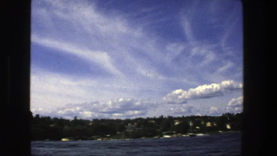 1975 Stockholm water view with a low wooded shore, rippled lake surface, distant buildings, and large cloud bands overhead.