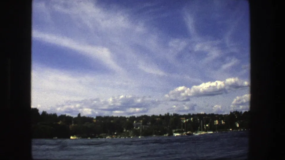 Stockholm waterfront view from 1975 showing choppy water, wooded shore, distant houses, sailboat masts, and a clear blue sky.