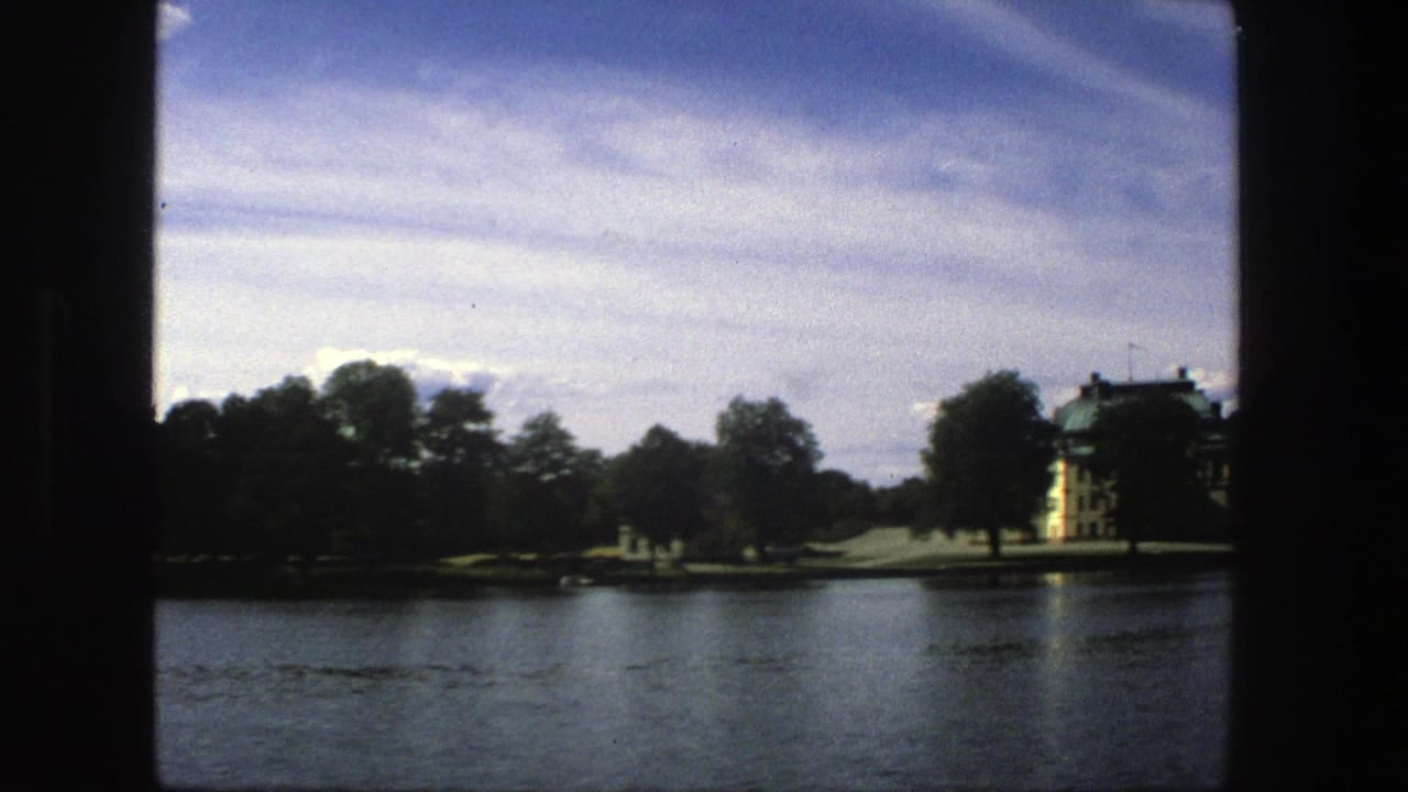 1975 Stockholm 8mm frame showing rippled water, a tree-lined shore and a yellow building with a green roof in the distance.