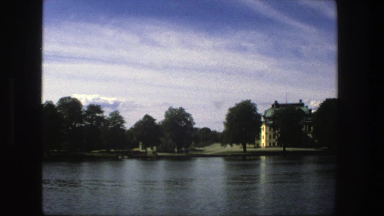 1975 Stockholm home-movie frame with a distant yellow waterfront building, trees, lawns and reflective lake water.