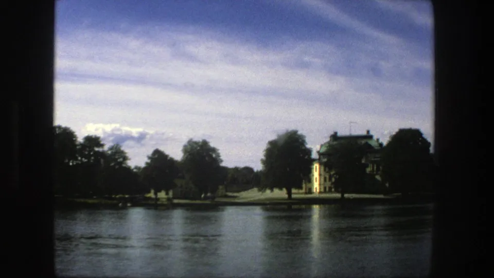 1975 Stockholm 8mm view across a lake, with the yellow building partly framed by trees and dark film edges.