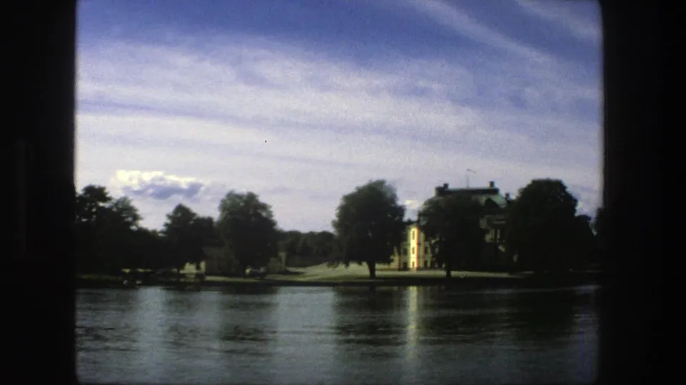 1975 Stockholm frame showing a yellow waterfront building, dark trees, open sky and a faint reflection on the lake.