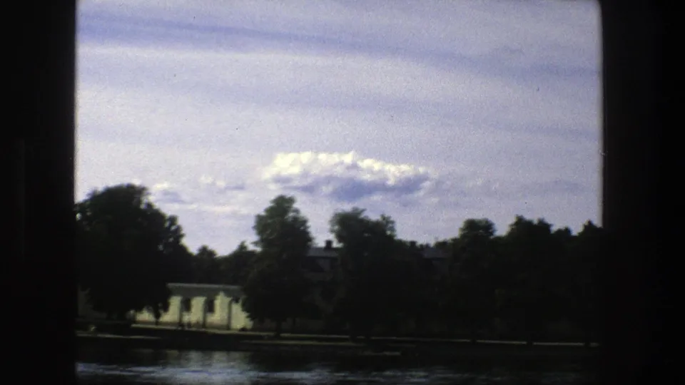 1975 Stockholm home-movie frame showing low pale buildings and trees across dark water beneath a clouded blue sky.