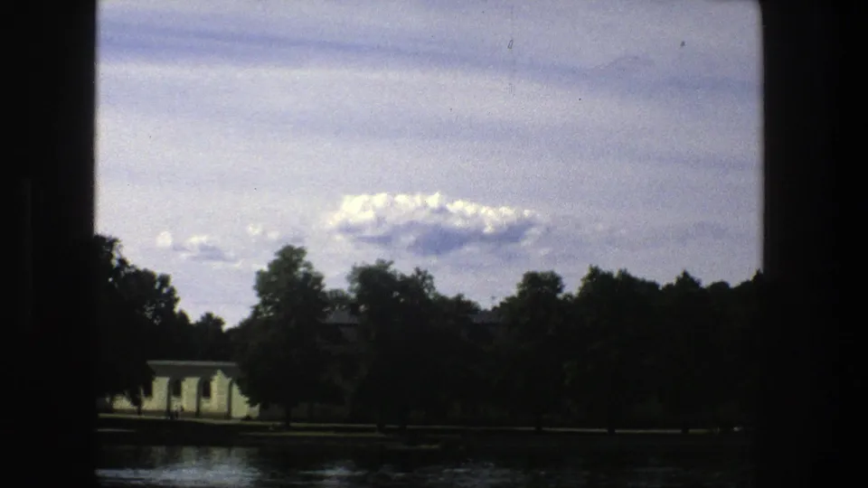 A tree-lined Stockholm waterfront with small pale buildings is seen across rippling water in a grainy 8mm frame.