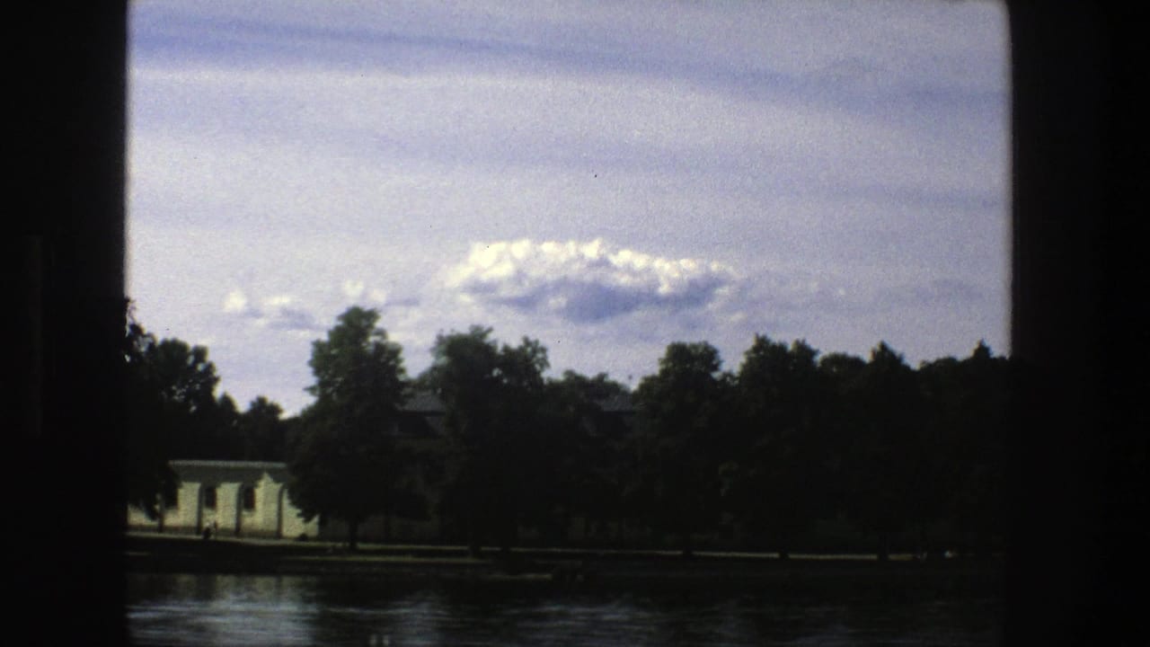 Cloudy sky over water, trees, and a pale building seen through dark film borders.