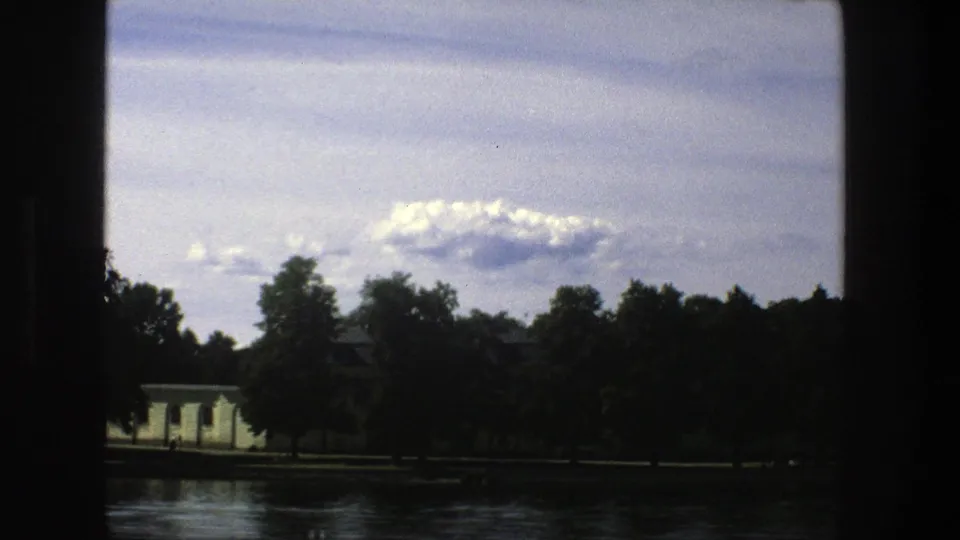 1975 Stockholm home-movie frame showing a calm waterfront, trees, cloudy sky, and a pale building through dark 8mm frame edges.