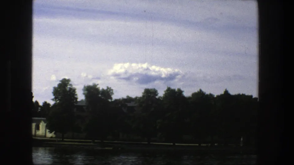 1975 Stockholm waterfront view with water, trees, a cloudy sky, and a pale building partially visible in the distance.