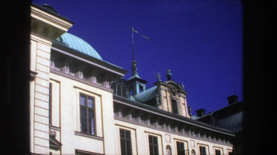 1975 Stockholm frame showing a classical building facade with green roof domes, tall windows, and a flagpole against deep blue sky.