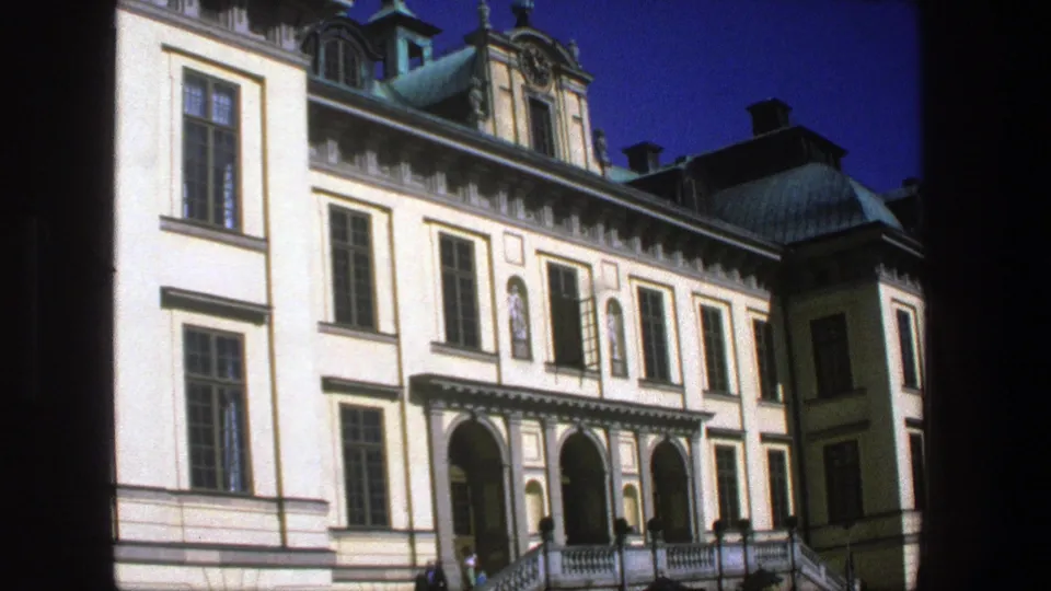 1975 Stockholm home-movie frame showing the lower part of a grand building, with arched doorways, exterior stairs, railings, and small pedestrians.