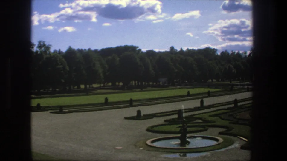 1975 Stockholm 8mm view across a formal garden with a central fountain, clipped hedges, broad paths, tree line, and scattered clouds.