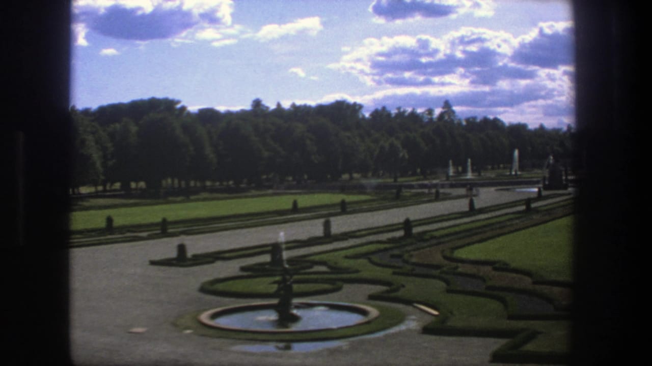 Formal garden with a round fountain, clipped hedges, lawns, paths, and trees under a cloudy sky.