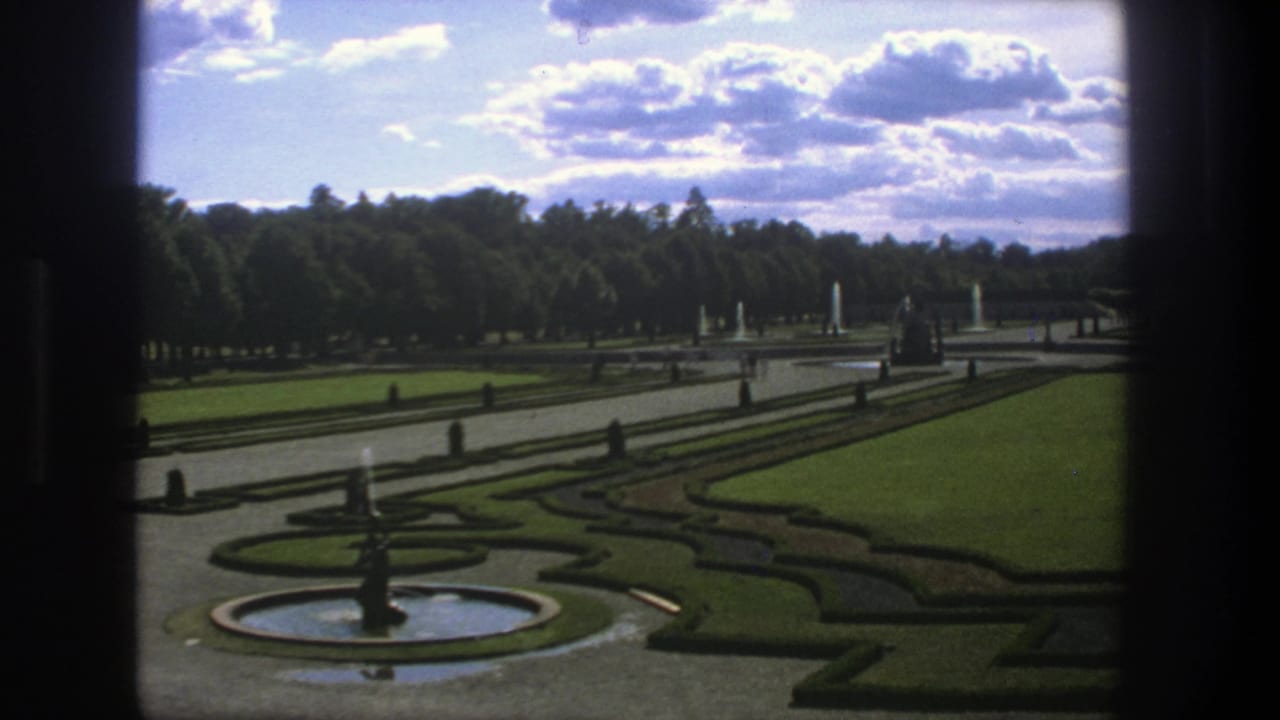Formal garden seen through dark vertical framing, with fountain, lawns, hedges, paths, and trees.