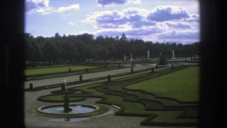 1975 Stockholm garden view with a central fountain, geometric hedges, green lawns, broad paths, distant tree line, and bright clouds.