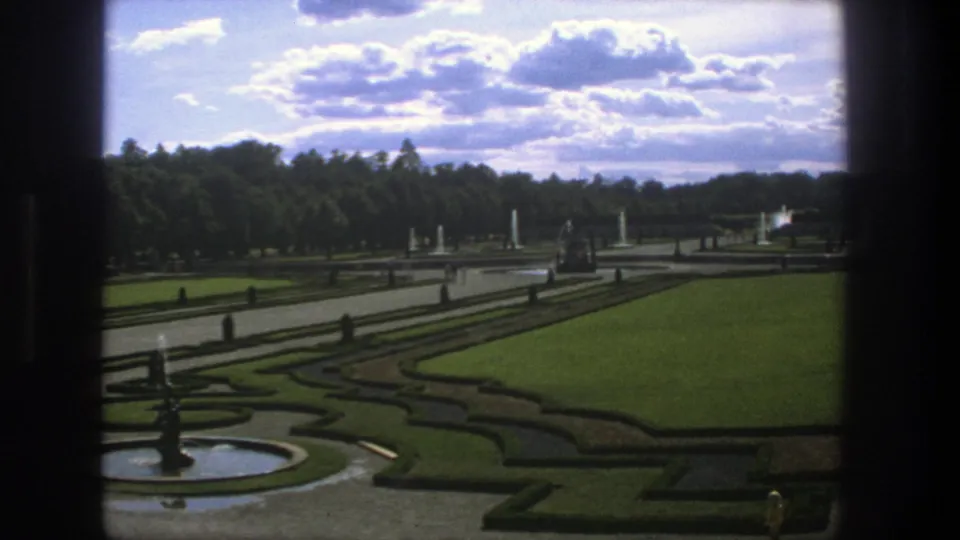 1975 Stockholm frame showing a formal garden after the pan, with foreground fountain basin, clipped hedge shapes, open lawn, paths, and trees.