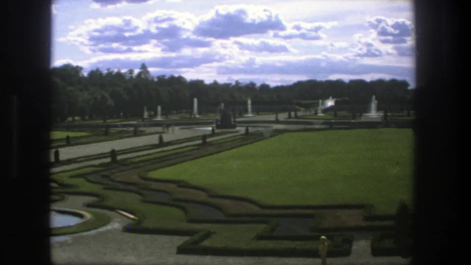 Late 1975 Stockholm garden frame with broad green lawn, clipped hedges, paved paths, several fountains, distant trees, and dark framing at the sides.