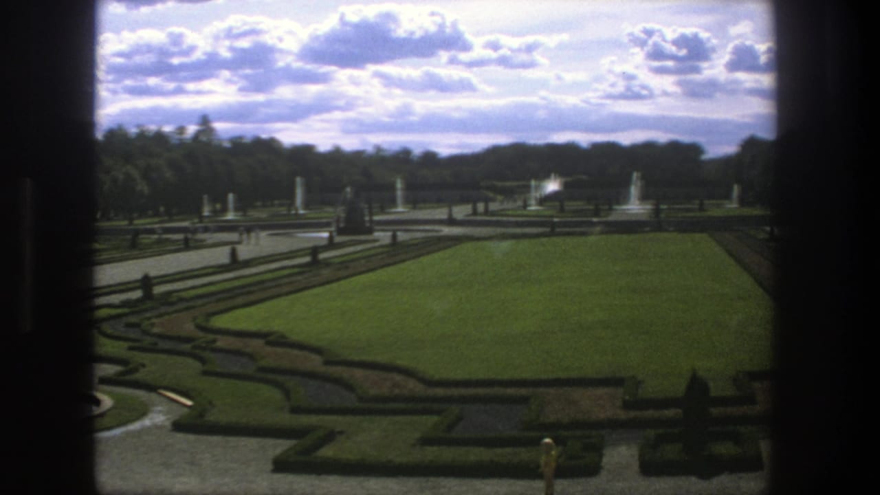 1975 Stockholm home-movie frame of a formal garden with clipped hedges, broad lawn, fountains, statues, trees, and dark window edges.