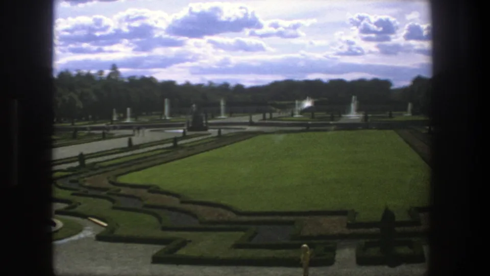 1975 Stockholm home-movie frame of a formal garden with clipped hedges, broad lawn, fountains, statues, trees, and dark window edges.