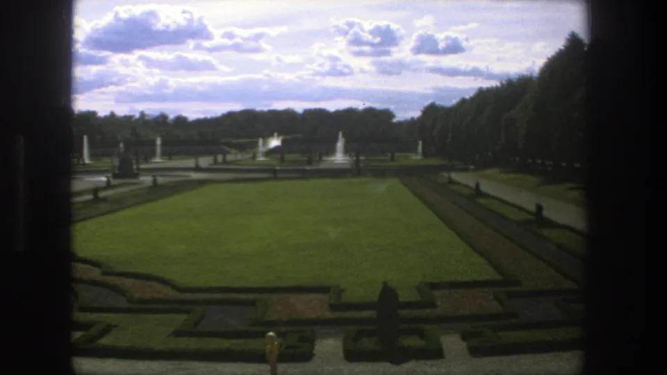 1975 Stockholm home-movie frame with a formal garden lawn, hedged beds, walkways, fountains, statues, and a dense tree row to the right.