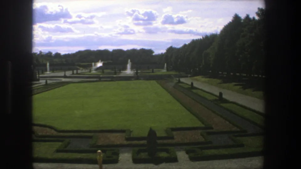 1975 Stockholm frame of a formal garden with large lawn, low hedges, fountains, statues, distant small figures, trees, and cloudy sky.